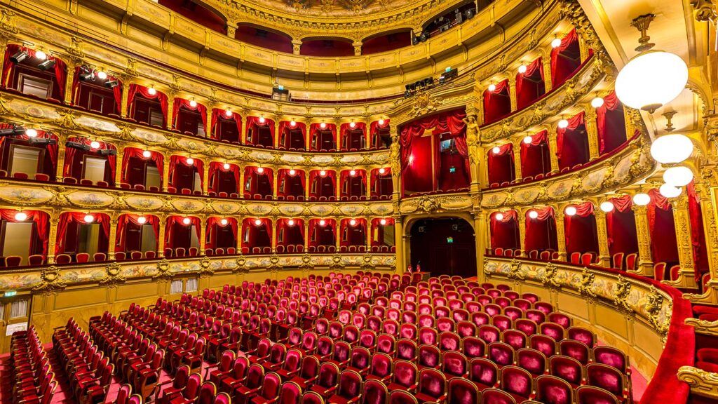Photo de la salle de spectacle de l'Opéra de Nice, vide. Fauteuils en velours rouge disposés en rangées, balcons aux ornements dorés.